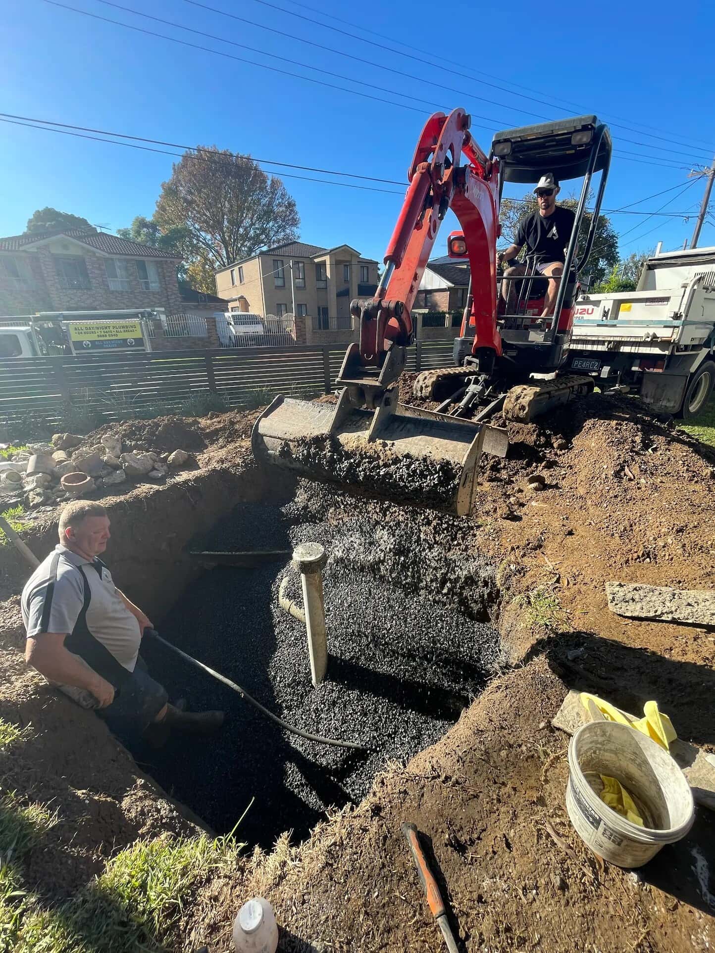 Excavator placing gravel around exposed sewer pipework in a yard trench