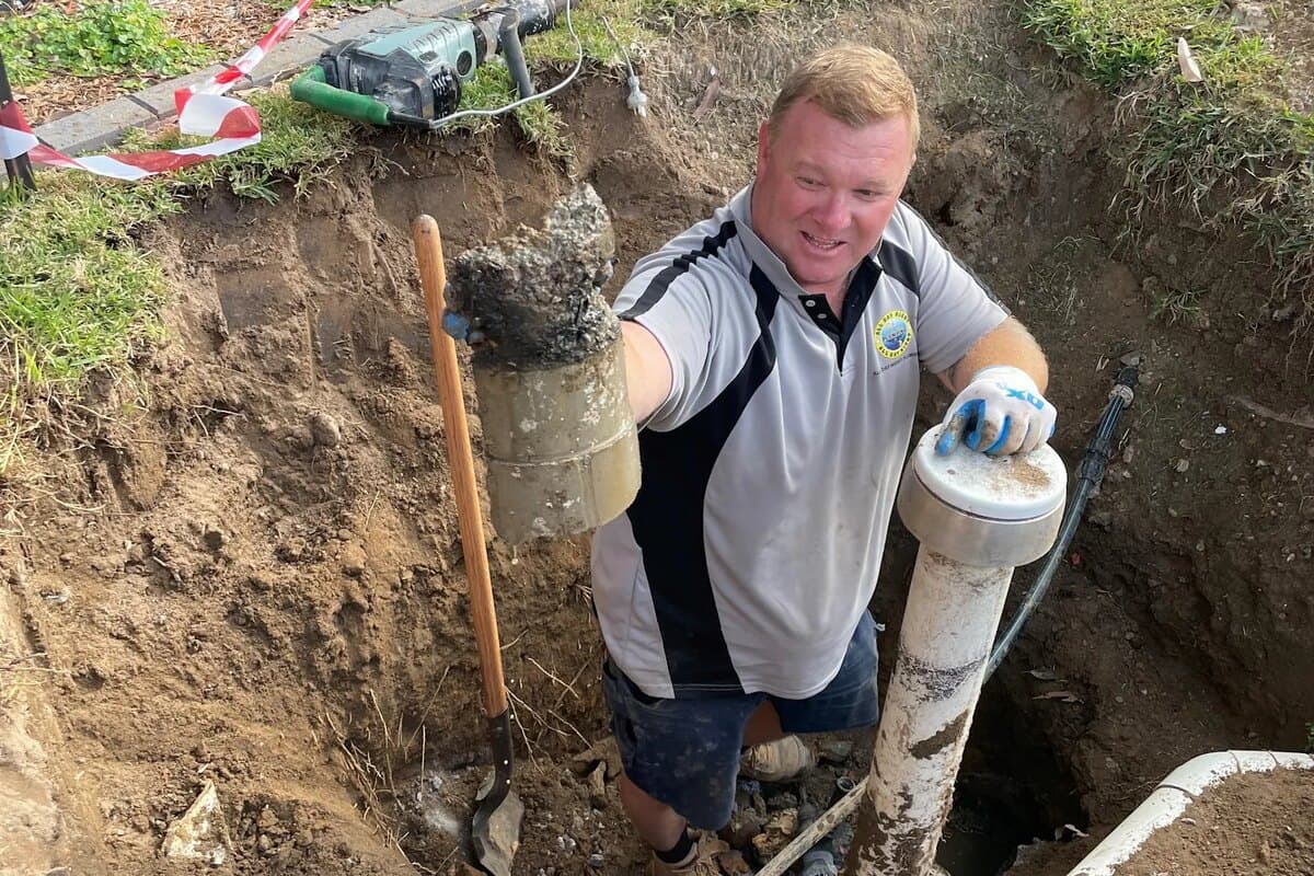 Scott Johnstone holding a concrete-obstructed sewer fitting in an excavated trench